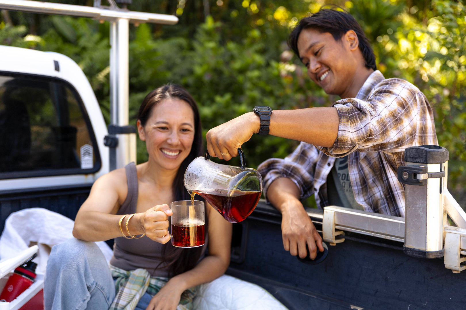 Man pouring a drink into a woman's mug in a vehicle's open back, surrounded by greenery.